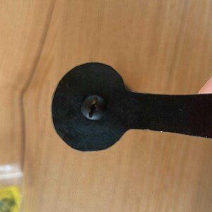 May include: Close-up of a black metal cabinet pull with a rounded end and a rectangular handle. The pull is attached with a single screw. The background is a light brown wooden surface.