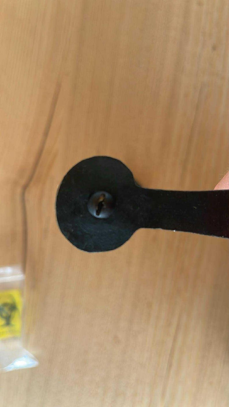 May include: Close-up of a black metal cabinet pull with a rounded end and a rectangular handle. The pull is attached with a single screw. The background is a light brown wooden surface.
