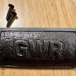 May include: A black cast iron drawer pull with the letters "GWR" embossed on the front. Two black screws are next to the pull. The pull has a textured surface and is on a wooden surface.