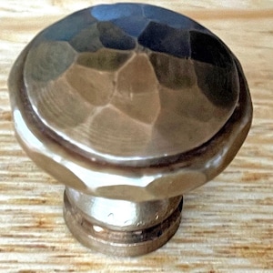 May include: A hammered metal cabinet knob with a rounded top and a cylindrical base. The knob has a textured surface with a dark, reflective finish. The base is a lighter, brushed metal.