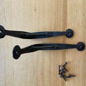 May include: Two black wrought iron gate hinges with diamond-shaped detailing and a set of screws. The hinges have a circular end and a curved handle. The items are on a light wood surface.