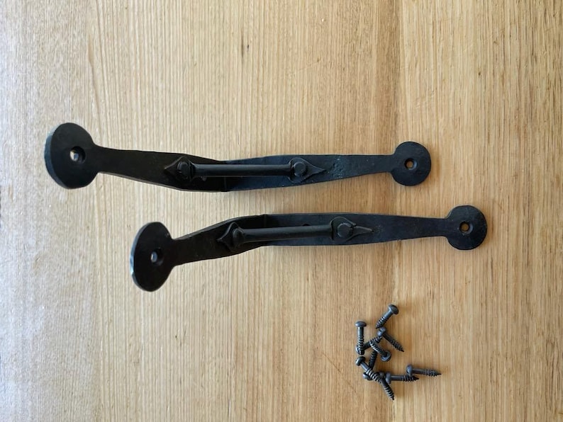 May include: Two black wrought iron gate hinges with diamond-shaped detailing and a set of screws. The hinges have a circular end and a curved handle. The items are on a light wood surface.