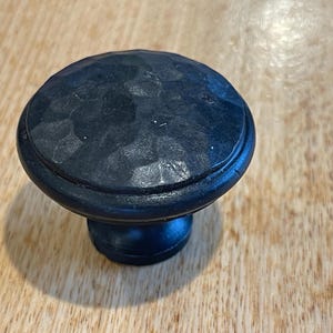 May include: A black, hammered metal cabinet knob with a rounded top and a short, cylindrical base. The knob has a textured surface and is set against a light brown wooden background. This hardware is suitable for drawers and cabinets.