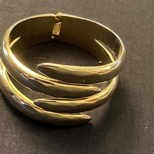May include: Gold-toned metal bracelet with a unique design. The bracelet features a hinged clasp and a layered, curved design that wraps around the wrist. The polished finish gives it a sleek, modern look.