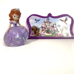 May include: A Sofia the First figurine wearing a purple glittery dress and a silver tiara, positioned next to a purple-framed decorative display featuring a printed illustration of a castle, a squirrel, a bird, and a rabbit in a garden setting.