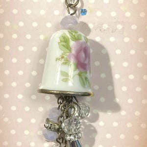 May include: A white porcelain thimble pendant with a pink rose design and gold trim. The thimble hangs from a chain with clear and lavender-colored beads and a silver-toned charm. The pendant is a unique piece of jewelry.