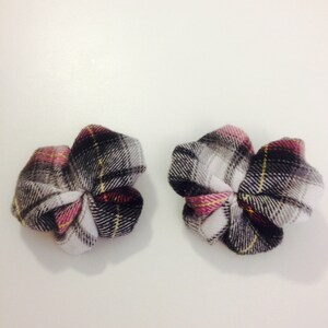 May include: Two fabric flower hair accessories. Each flower is made of a plaid pattern in black, white, gray, pink, and yellow. The flowers are arranged on a white surface.