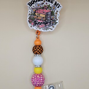 Könnte beinhalten: Ein Ausweis-Rollhalter mit weißem und floralem Design und dem Text "Books are my Therapy". Die Rolle hat ein Perlendesign mit orangefarbenen, schwarz-weiß karierten, weißen, gelben und rosa Strassperlen. Ein durchsichtiger Kunststoffclip ist angebracht.