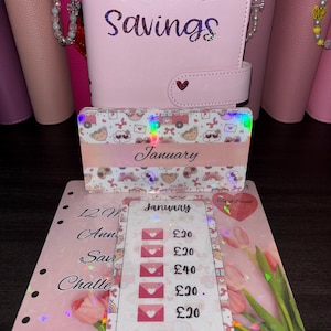 May include: A light pink binder with the text Annual Savings in dark lettering and a heart-shaped snap closure. In front are two rectangular cards featuring a January label, a pattern of small icons, and a savings tracker with £20 increments.