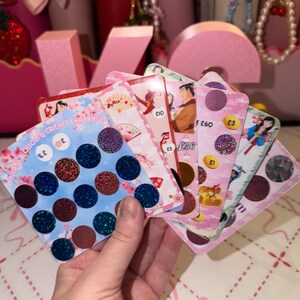 May include: A collection of Mulan-themed savings challenge cards featuring various illustrations of characters and fans. The cards include circular scratch-off areas with holographic textures, printed currency symbols, and numerical values like £10, £5, £60, and £3.