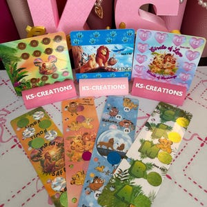 May include: A collection of seven rectangular savings challenge cards featuring Lion King characters, colourful illustrations, and scratch-off circles with currency values. Each card displays the text KS-CREATIONS, Simba, Timon, Pumbaa, and various savings goals in pounds.