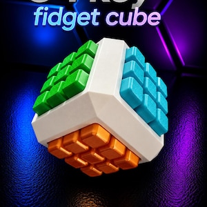May include: A 54 key fidget cube with a white frame and three distinct sides featuring green, blue, and orange square buttons. The text 54 key fidget cube is displayed in white and purple lettering above the object.