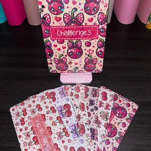 May include: A set of planner inserts featuring a repeating pattern of pink and red cherries with bows, hearts, and sparkles. The top sheet has a rectangular label with the text "Challenges" and the bottom sheet has a label reading "Challenge one".