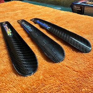 May include: Three black carbon fiber shoe horns with the text "PureBike" in iridescent lettering. The shoe horns are arranged on an orange towel. The carbon fiber has a woven pattern.