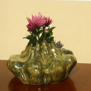 May include: A green and gold ceramic vase with a distinctive, textured design, holding a cluster of pink chrysanthemums. The vase has an organic shape and sits on a dark wooden surface, creating a striking contrast.