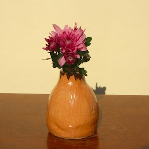 May include: A small, orange ceramic vase with a textured surface holds a cluster of vibrant pink flowers. The vase sits on a dark brown wooden surface, with a light yellow wall in the background. The flowers have dark green leaves.
