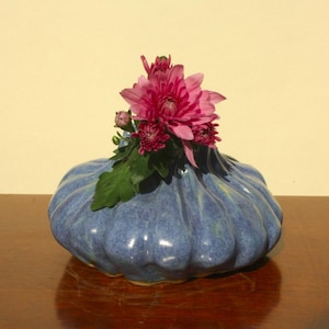 May include: A blue ceramic vase with a fluted design, holding a cluster of pink flowers and green leaves. The vase sits on a dark wooden surface, with a light-coloured wall in the background.