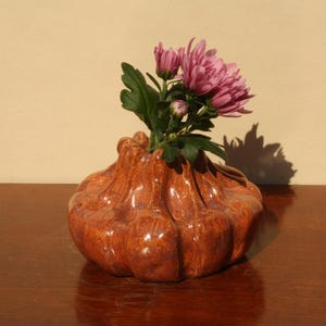 Könnte beinhalten: Eine braune Keramikvase mit bauchiger Form, die eine Gruppe rosa Chrysanthemen enthält. Die Vase hat eine glänzende Oberfläche und steht auf einer dunklen Holzoberfläche. Die Blumen haben grüne Blätter.