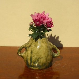 May include: A handmade, green ceramic vase with two handles, containing a bouquet of pink flowers. The vase has a glossy finish and is on a wooden surface. The background is a light-coloured wall.