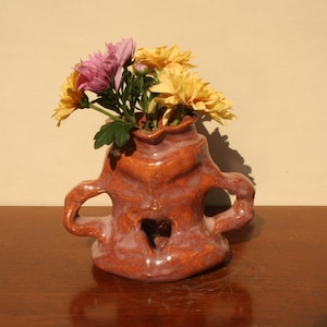 Könnte beinhalten: Eine handgefertigte Keramikvase in Orange- und Rottönen, mit herzförmigem Ausschnitt und zwei Henkeln. Sie enthält einen Strauß gelber und violetter Chrysanthemen. Die Vase steht auf einer Holzoberfläche. Die Vase ist etwa 15 cm hoch.