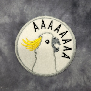 May include: Embroidered patch with a white cockatoo design, yellow crest, and open beak, with the word "AAAAA" in black. The patch has a grey border and is on a grey background.