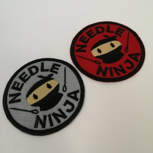 May include: Two circular embroidered patches. One is grey with black text and a ninja face, the other is red with black text and a ninja face. Both patches read "NEEDLE NINJA" around the edge.