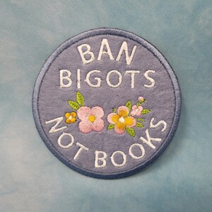 May include: A circular embroidered patch in blue with the text "BAN BIGOTS NOT BOOKS" in white. The patch is decorated with floral embroidery in pink, yellow, and green. The design is a statement piece.