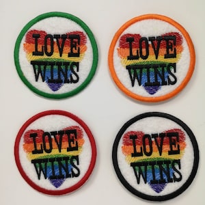 May include: Four round embroidered patches with the words "LOVE WINS" in black. Each patch features a rainbow heart design. The patches have different colored borders: green, orange, red, and black. The background is white.