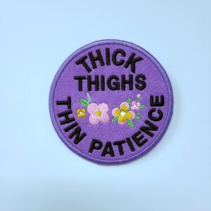 May include: A round, purple embroidered patch with the words "THICK THIGHS THIN PATIENCE" in black. The patch features embroidered flowers in pink, yellow, and white with green leaves. The patch is on a white background.