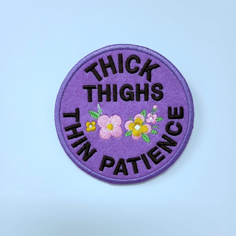 May include: A round, purple embroidered patch with the words "THICK THIGHS THIN PATIENCE" in black. The patch features embroidered flowers in pink, yellow, and white with green leaves. The patch is on a white background.
