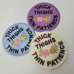 Könnte beinhalten: Drei runde, gestickte Aufnäher in Hellblau, Lila und Creme. Jeder Aufnäher trägt den Text "THICK THIGHS THIN PATIENCE" und florale Stickereien in Rosa und Gelb.