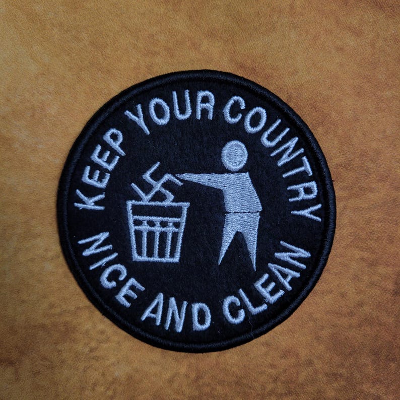 May include: A round, black embroidered patch with white text that reads "KEEP YOUR COUNTRY NICE AND CLEAN." The patch features a graphic of a person throwing a swastika into a trash can.