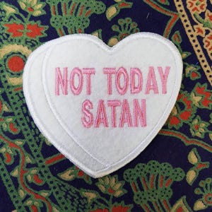 May include: White heart-shaped patch with pink embroidered text reading "NOT TODAY SATAN." The patch has a white border and is set against a patterned fabric background with blue, green, and red hues.