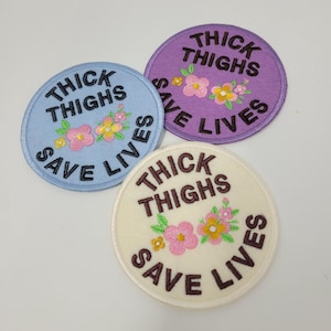 May include: Three embroidered patches in light blue, lavender, and cream. Each patch features the text "THICK THIGHS SAVE LIVES" in a circular design, with a floral arrangement in the center. The patches are circular or oval.