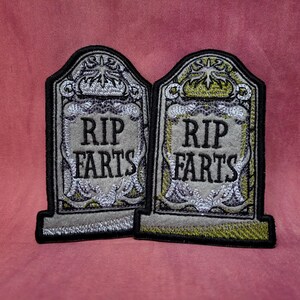 Peut inclure: Deux écussons brodés en forme de pierres tombales. Les écussons sont gris avec des bordures noires et comportent le texte "RIP FARTS" en noir. Le design comprend des accents décoratifs argentés et dorés, créant un thème gothique humoristique.