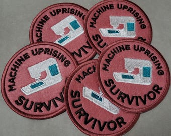 Puede incluir: Un conjunto de parches redondos bordados. Cada parche es de color rosa polvoriento con las palabras "MACHINE UPRISING" y "SURVIVOR" en negro. Una máquina de coser blanca con detalles turquesa está en el centro.