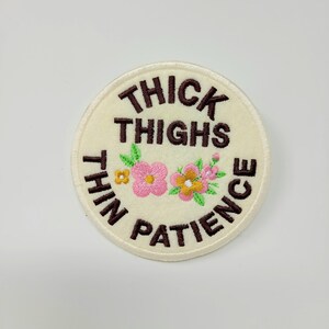 May include: A round, cream-colored embroidered patch with the text "THICK THIGHS THIN PATIENCE" in brown. The patch features embroidered pink and yellow flowers with green leaves.
