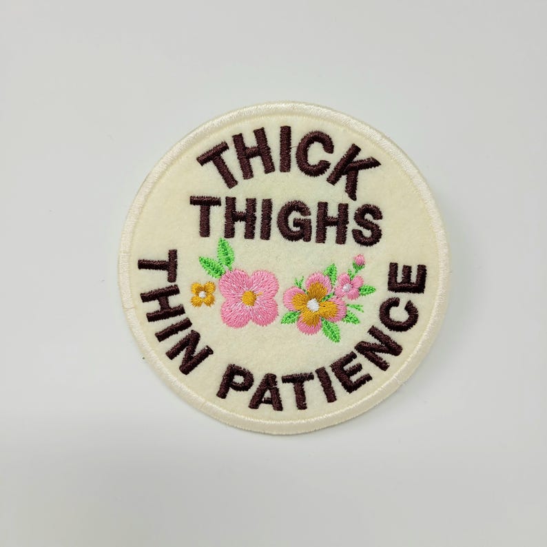 May include: A round, cream-colored embroidered patch with the text "THICK THIGHS THIN PATIENCE" in brown. The patch features embroidered pink and yellow flowers with green leaves.