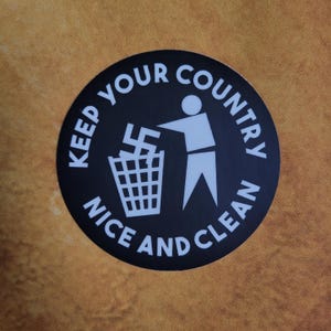 May include: A round black sticker with white text and graphics. The text reads "KEEP YOUR COUNTRY NICE AND CLEAN." A person figure is throwing a swastika into a trash can. The background is a textured, golden-brown color.