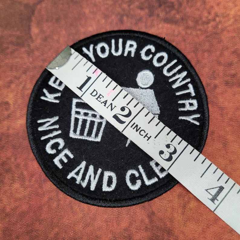 May include: A round, black patch with white embroidered text that reads "KEEP YOUR COUNTRY NICE AND CLEAN." The patch features a graphic of a person throwing trash into a bin. The patch is approximately 2 inches in diameter.