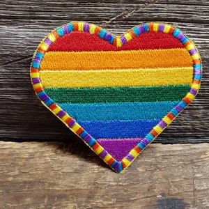 Puede incluir: Parche bordado en forma de corazón con diseño de arcoíris. El parche presenta rayas rojas, naranjas, amarillas, verdes, azules y moradas, y un borde colorido. Se encuentra sobre un fondo de madera rústica. Ideal para chaquetas, bolsos u otros artículos de tela.