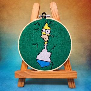 May include: Embroidered artwork in a hoop, depicting Homer Simpson emerging from green shrubbery. The design shows Homer's yellow skin, white shirt, blue trousers, and distinctive expression. The artwork is displayed on a small wooden easel.