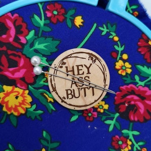 Könnte beinhalten: Ein blauer Stickrahmen hält Stoff mit einem Blumenmuster in Rot, Rosa, Gelb und Grün. Ein Holz-Kreis mit dem Text "HEY ASS BUTT" befindet sich in der Mitte, mit zwei perlenbesetzten Nadeln.