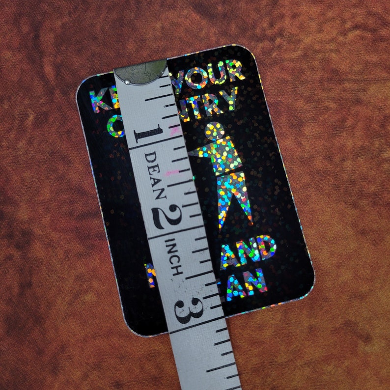 May include: A black rectangular sticker with holographic text and a figure. The text reads "KEEP YOUR ENTRY AND FAN". The sticker is approximately 2 inches (5 cm) in height and is being measured with a white measuring tape.