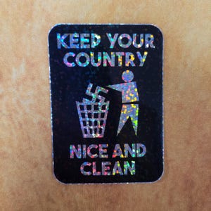 May include: A rectangular sticker with a black background and holographic glitter effect. The sticker features the text "KEEP YOUR COUNTRY NICE AND CLEAN" above an illustration of a person throwing a symbol into a trash can.