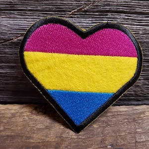 Könnte beinhalten: Ein herzförmiger Aufnäher mit schwarzem Rand, der ein Design in Rosa, Gelb und Blau aufweist. Der Aufnäher ist wahrscheinlich ein gesticktes Emblem, möglicherweise für Kleidung oder Accessoires. Die Farben sind in horizontalen Streifen angeordnet.
