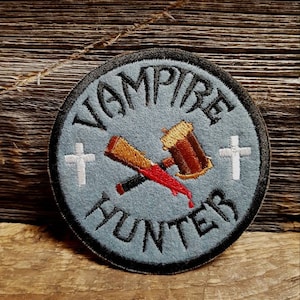 May include: Embroidered patch with the words "VAMPIRE HUNTER" in black lettering on a light blue background. The patch features a hammer, stake, and two white crosses. The patch has a black border and is set against a wooden background.