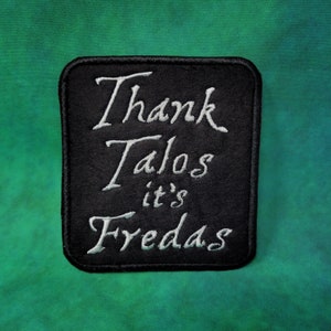 Può includere: Toppa quadrata nera con testo ricamato bianco che recita "Thank Talos it's Fredas". La toppa ha angoli arrotondati e un bordo nero. Lo sfondo è un gradiente verde e blu.