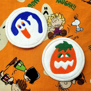 May include: Two circular, white embroidered patches with Halloween designs. One shows a blue and white ghost with an orange mouth, the other an orange jack-o'-lantern with a green stem. The background is orange with cartoon characters.