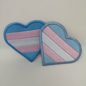 May include: Two heart-shaped patches with the transgender flag design. Each patch features a light blue border and a light blue background with pink and white stripes. The patches are embroidered.